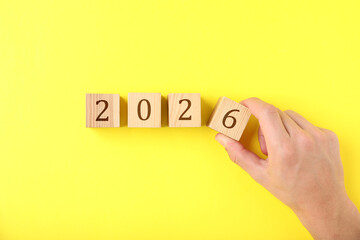 New 2026 year. Man putting cube with number 6 on yellow background, top view