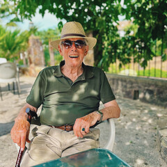 Senior man with straw hat and brown sunglasses in green polo shirt sits in chair of outdoor terrace of cafe.