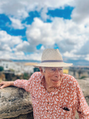 Senior man with straw hat in red-white shirt stands by a stone wall of an old bridge under blue sky with clouds.