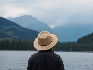 Woman in straw hat gazes at serene lake and misty blue mountains, embracing tranquil nature and peaceful outdoor adventure