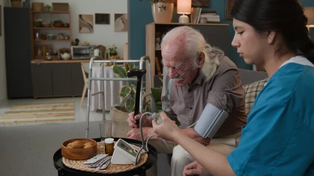 Side view shot of young woman as female nurse visiting elderly man at home and checking blood pressure using manual pump, copy space