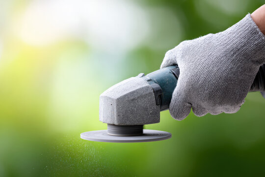 Close up of gloved hands using cordless grinder to sharpen metal with green blurred background and sunlight