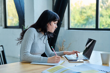 Young woman university student sitting at desk with laptop, typing writing studying