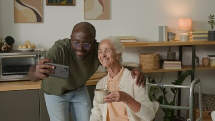 Medium shot of Black man as social worker taking selfie with white haired woman at home both smiling looking at smartphone camera, copy space - Powered by Adobe