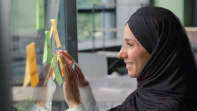 Smiling Muslim business woman stick sticky notes on glass in modern office Arabian businesswoman in hijab preparing planning work tasks female girl corporate leader leaving note organize job strategy