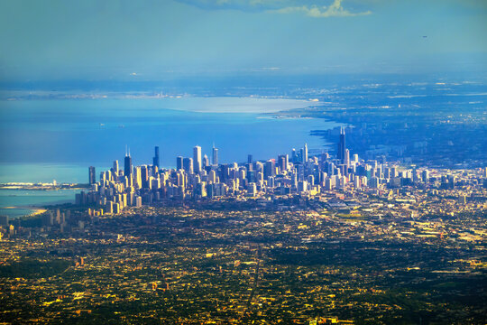 Aerial view of Chicago skyline by Lake Michigan: dense high-rises, waterfront, surrounding suburbs and green spaces in low-angle sunlight.