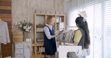 Two asian female fashion designers analyzing dress detail on clothing rack together while using digital tablet for reference in modern tailoring studio with natural light and stylish workspace