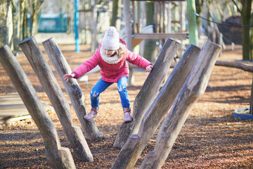 A young girl in warm winter clothing balancing and climbing on wooden playground equipment outdoors