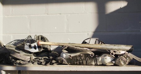 Sitting stacked catcher's gear on metal bench at dugout, chest pads, shin pads, masks, bat, straps