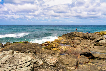 Volcanic coastline near the Bañaderos in Arucas, Gran Canaria: strong Atlantic waves hit the dark lava rocks, while people relax or explore the rugged natural setting.
