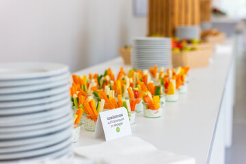 Close-up of an elegantly arranged buffet table with a white tablecloth. Glasses filled with vegetable Sticks, with slightly blurred fresh fruit in the background.