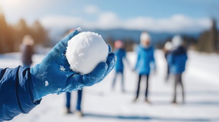 Close-up of hand in blue winter gloves holding round snowball outdoors. Snowy field and kids playing in soft blurred background. Winter fun and seasonal outdoor activity concept with copy space