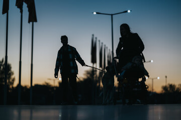 Fototapeta premium Silhouettes of a family walking at sunset: a man with a dog on a leash and a woman with a stroller, creating a peaceful, everyday moment outdoors.