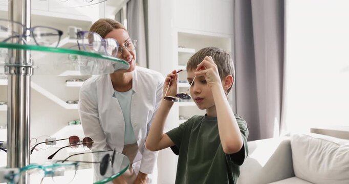 Young female optician helping to little boy with sunglasses selection
