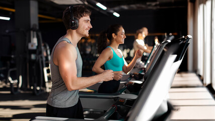 Sporty smiling man running on treadmill in gym, wearing headphones, enjoying morning workout with music, side view, panorama