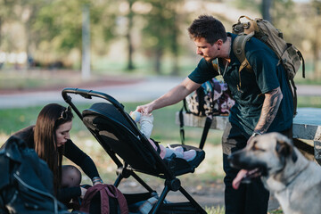 A family enjoys a day at the park: a dad with a backpack feeds a baby in a stroller while a mother checks a bag nearby, with a dog nearby and calm, sunny surroundings.