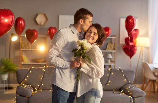 Happy loving couple enjoying Valentine Day or anniversary at home. Smiling woman holding bouquet of white roses as man kissing her forehead in room filled with heart shaped balloons and fairy lights.