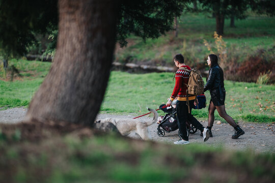 Couple strolls through a green park with a baby stroller and a leashed dog. The man wears a red sweater and backpack, while the woman in a black jacket walks beside him, enjoying outdoor family time.