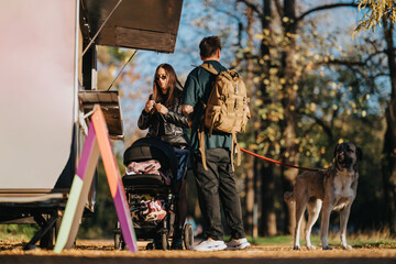 A couple with a backpack chats near a colorful food truck in a sunlit autumn park. A dog on a leash stands nearby as a stroller rests beside them, capturing a relaxed family moment.