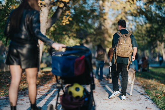 A man wearing a backpack walks a dog along a sunlit autumn park path while a woman pushes a stroller in the distance. Golden leaves scatter the ground, creating a warm, family-friendly moment.