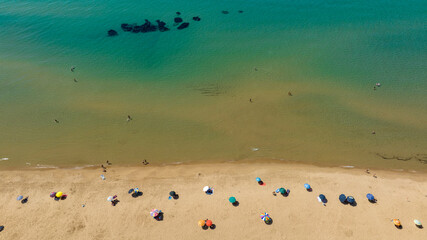 Perpendicular aerial view of a sandy beach overlooking the emerald-green Mediterranean Sea. A few people on vacation with their umbrellas on the coast.