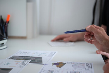 Zwei Personen besprechen technische Zeichnungen und Pl&auml;ne an einem wei&szlig;en Schreibtisch, Fokus auf Hand mit Kugelschreiber und die Dokumente, moderne B&uuml;roatmosph&auml;re.