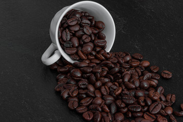 Overturnerd ceramic cup with coffee beans on a dark surface.