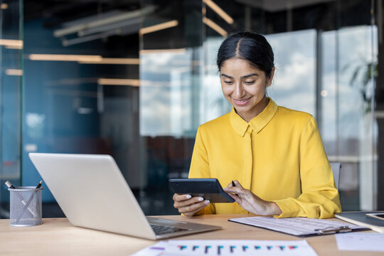 Young smiling Indian woman working in the office, sitting at a desk with a laptop and documents, using a calculator