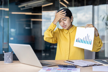 Young smiling Indian woman working in the office, sitting at a desk with a laptop and documents, using a calculator
