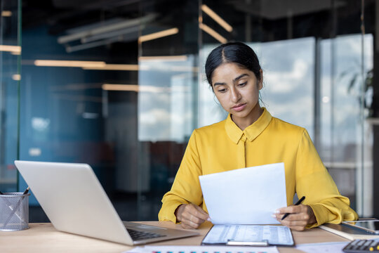 Serious and confused young Indian woman sitting in the office at a table with a laptop, working with documents and data
