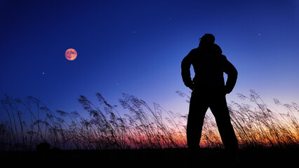 Silhouette of a man and countryside under the Moon, stars and planets in blue hour time.