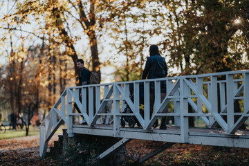 A family-friendly scene on a white wooden bridge in a sunlit autumn park. People walk, a stroller is pushed, leaves fall around, creating a warm, relaxed outdoor moment.