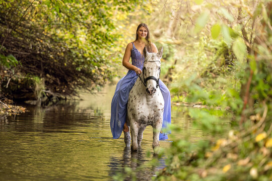 Young woman in a flowing blue dress standing beside her Danish Knabstrupper gelding while both stand peacefully in a calm forest river.