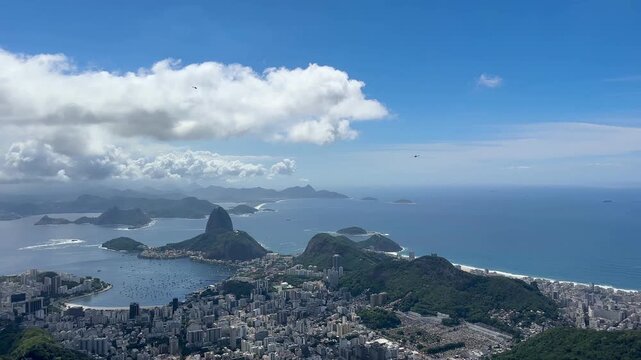 Panoramic view of Rio de Janeiro from Corcovado, featuring Sugarloaf Mountain, Guanabara Bay, Santos Dumont Airport, and the Atlantic coastline under bright, partly cloudy weather.