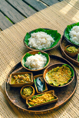 Traditional Thai food is served in wooden bowls with round and square shapes on a bamboo tray with spoons. There is plain rice, omelet, curry, and chili paste on a woven mat with soft sunlight.