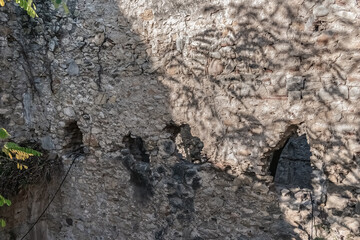 Weathered ancient stone wall in Jardins dels Alemanys, Girona, Spain, featuring historic fortification masonry, small openings, and textured surfaces lit by dappled sunlight