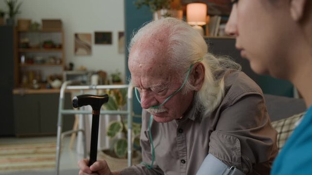 Tilt up shot young woman as female nurse checking blood pressure and caring for senior man at home with focus flowing from hands using manual pump to face of patient