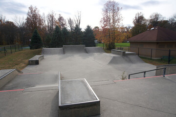 Views of an empty, outdoor concrete skate park featuring various ramps, quarter pipes, ledges, and funboxes, set within a park landscape with trees showing late autumn foliage. Some sections of the pa