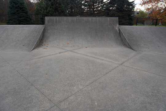 Views of an empty, outdoor concrete skate park featuring various ramps, quarter pipes, ledges, and funboxes, set within a park landscape with trees showing late autumn foliage. Some sections of the pa