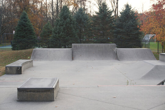 Views of an empty, outdoor concrete skate park featuring various ramps, quarter pipes, ledges, and funboxes, set within a park landscape with trees showing late autumn foliage. Some sections of the pa