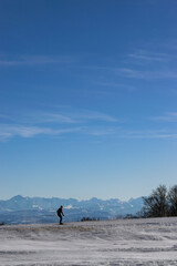Obraz premium Lone cross-country skier gliding across snowy plateau with panoramic view of distant alpine mountains under clear blue sky