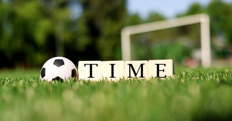 A soccer ball rests on vibrant green grass beside wooden blocks arranged to spell out the word time, while a goal post stands in the background under bright daylight