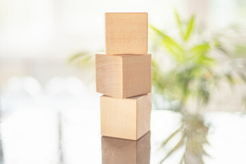Three simple wooden blocks are stacked vertically on a shiny surface. Soft light shines in from a nearby window, creating a calm atmosphere with greenery in the background