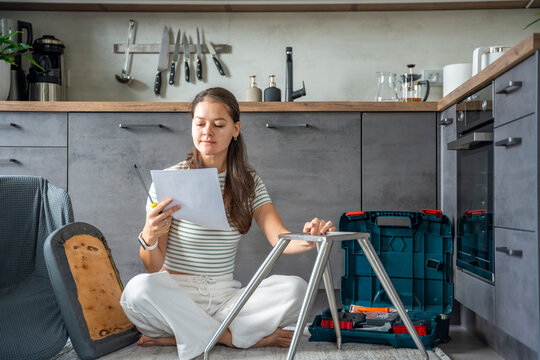 Smiling young woman sitting on the floor reading instructions while assembling a chair. Continuation of the series showing joy and satisfaction from independent DIY work at home.