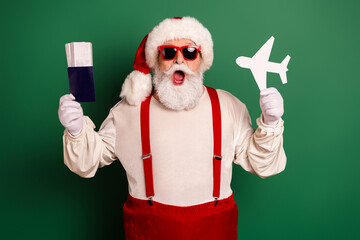 Santa in red suspenders and a white beard with red glasses holds a passport and paper plane in a festive Christmas scene against a green background