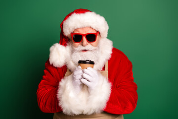 Santa in a red suit with hat and white beard holds a coffee cup with gloved hands against a green background for holiday season mood