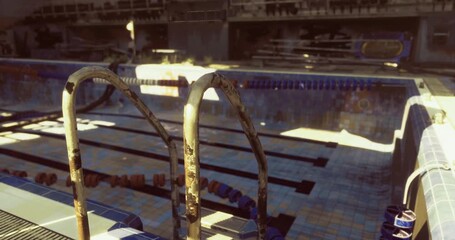 An abandoned swimming pool area has a rusted ladder leading into the water. Sunlight reflects off the blue tiles, highlighting the empty lanes with unused diving boards in the background.