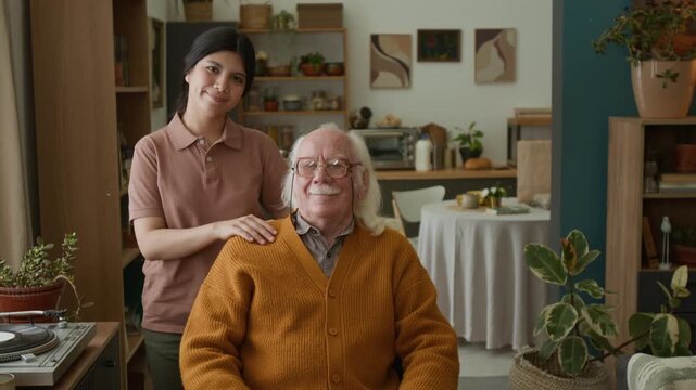 Medium shot of young woman posing with senior man in wheelchair looking at each other then looking at camera with smiles, copy space