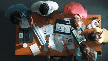 Top down of skilled business people holding tablet an looking at blueprint. Aerial view of architect engineer planning and design house design together while standing at table with laptop. Symposium.