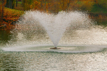 Water fountain creates a beautiful spray in the tranquil lake surrounded by autumn foliage during a sunny afternoon